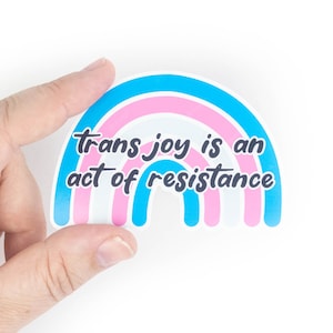 以下が含まれることがあります： 青、ピンク、白のレインボーデザインのステッカー。ステッカーには「trans joy is an act of resistance」という黒いテキストが書かれています。