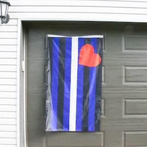 May include: A blue, black, and white striped flag with a red heart in the center. The flag is hanging on a garage door.