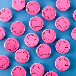 May include: A collection of pink round buttons with black outlines of t-shirts on them.