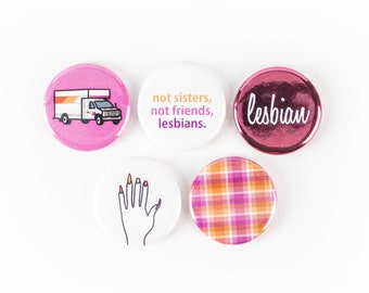 May include: Five pink and white button pins with different designs. One pin features a pink and orange plaid pattern. Another pin features a white hand with pink and orange nail polish. A third pin features a white moving truck on a pink background. A fourth pin features the text "lesbian" in white on a pink background. The last pin features the text "not sisters, not friends, lesbians." in pink and white on a white background.