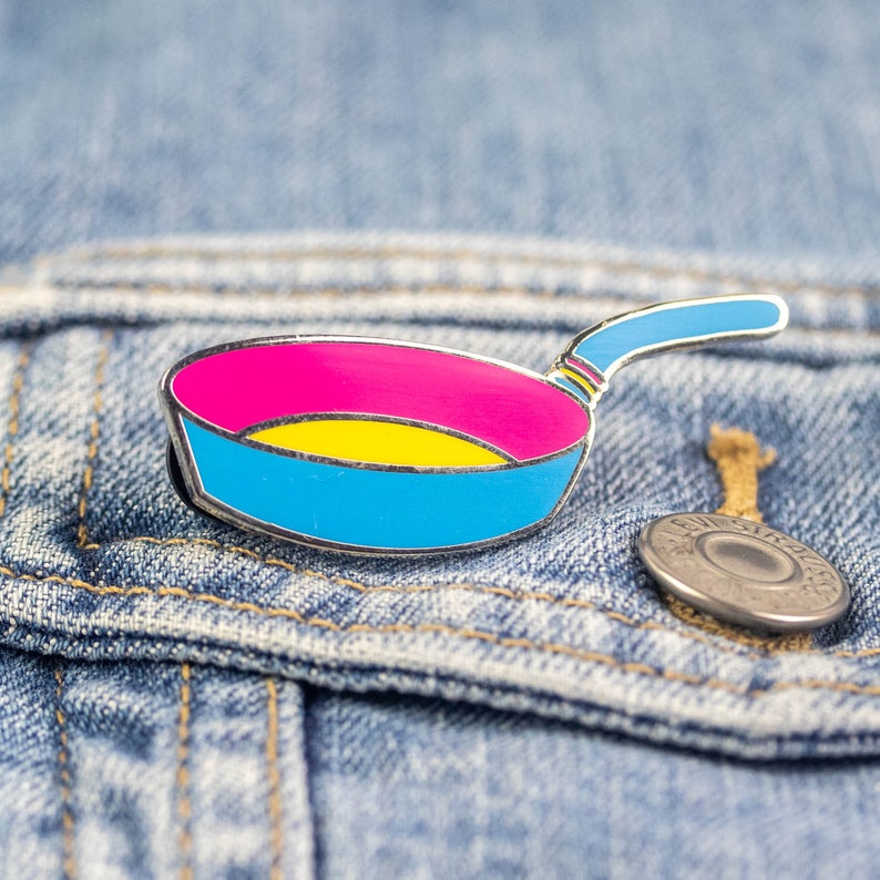 Puede incluir: Un pin de esmalte plateado con forma de sart&eacute;n con un dise&ntilde;o azul, amarillo y rosa. El pin est&aacute; sobre un fondo de mezclilla azul.
