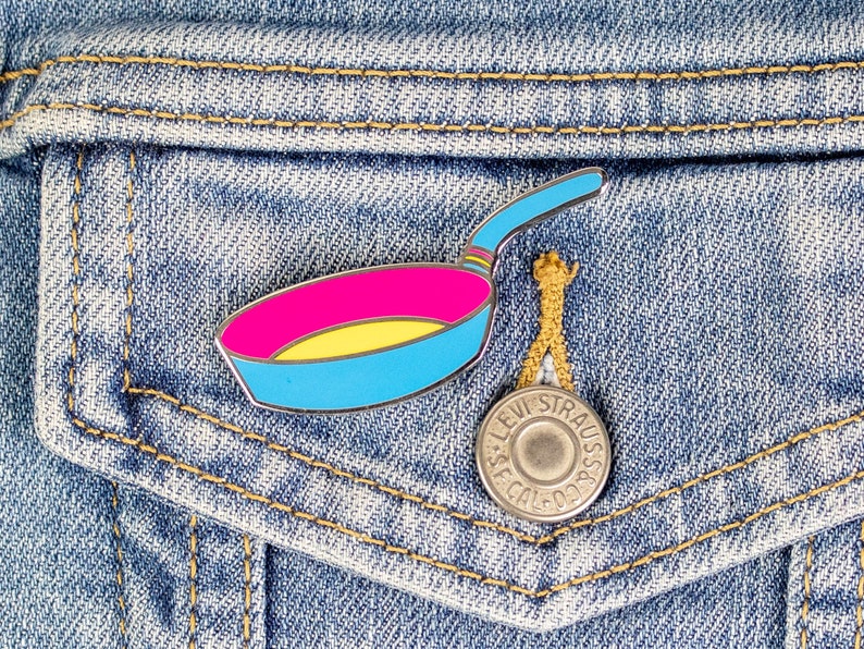 Puede incluir: Un pin de esmalte azul, amarillo y rosa con forma de sart&eacute;n. El pin est&aacute; sujeto a una chaqueta vaquera.