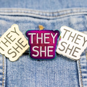 May include: Three enamel pins with the text "They She" on a blue denim background. The pins are gold, silver, and iridescent.
