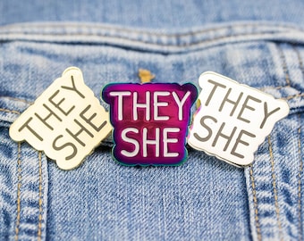 May include: Three enamel pins with the text "They She" on a blue denim background. The pins are gold, silver, and iridescent.