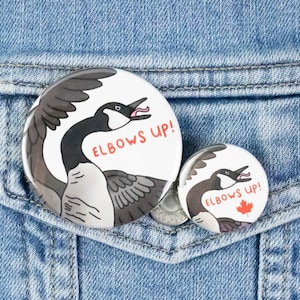 Puede incluir: Dos botones redondos con un diseño de ganso de dibujos animados. El botón más grande tiene el texto "Elbows Up!" en rojo. El botón más pequeño tiene el mismo diseño y texto, pero es más pequeño. Los botones están sobre un fondo de mezclilla azul.