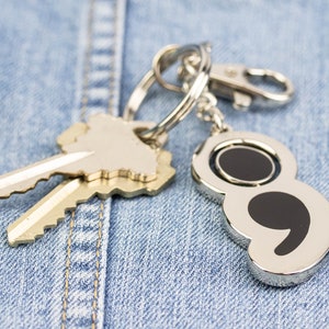 May include: A silver key ring with two keys and a silver charm shaped like a semicolon. The keyring is hanging on a blue denim background.