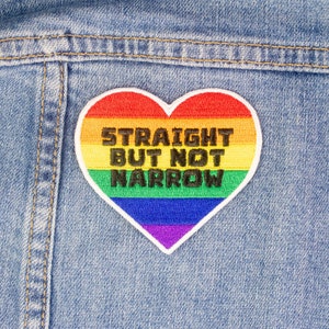 Puede incluir: Un parche en forma de corazón arcoíris con el texto "Straight But Not Narrow" cosido en una chaqueta vaquera azul.