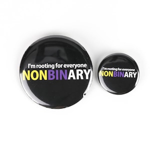 Puede incluir: Dos pines de botón negros con el texto "I'm rooting for everyone NONBINARY" en letras blancas con acentos amarillos y morados. El pin más grande tiene un diámetro de aproximadamente 5 cm y el pin más pequeño tiene un diámetro de aproximadamente 2,5 cm.