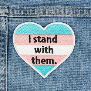 May include: A heart-shaped patch with the transgender pride flag colors and the text "I stand with them."