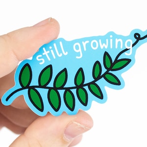 Könnte beinhalten: Ein hellblauer Aufkleber in Form eines Blattes mit den Worten "still growing" in Weiß. Das Blatt hat grüne Blätter mit schwarzen Umrissen. Der Aufkleber wird in der Hand einer Person gehalten.