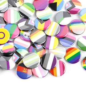 May include: A collection of colourful round buttons featuring various pride flags and symbols. The buttons are made of metal and have a shiny finish.