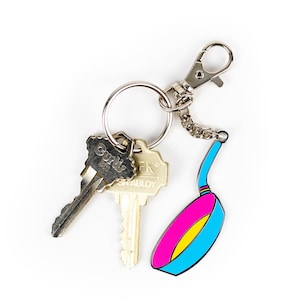 May include: A silver key chain with two keys and a blue and pink frying pan charm. The keys have the words "Curly" and "K's" on them.