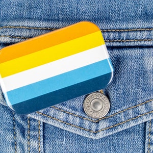 May include: A colorful pin with stripes of yellow, orange, white, blue, and dark blue on a denim jacket.