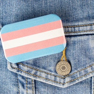 May include: A blue denim jacket with a transgender pride flag pin on the pocket.