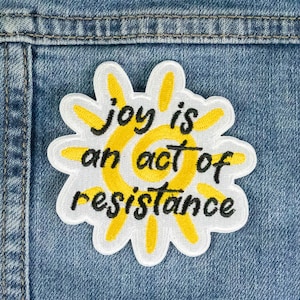 Könnte beinhalten: Ein weißer und gelber gestickter Aufnäher mit einem Sonnenmotiv und dem Text "joy is an act of resistance".