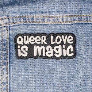 May include: A black and white embroidered patch on a blue denim background. The patch says "Queer Love is Magic".