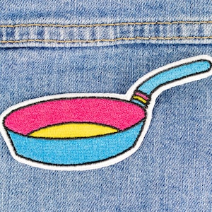 Op de afbeelding: Een kleurrijk geborduurd pleister van een koekenpan op een blauwe denim achtergrond. De pan is wit omrand, met een blauwe steel, een gele binnenkant en een roze rand.