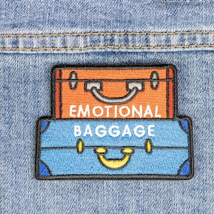Puede incluir: Un parche bordado azul y naranja con el texto "Emotional Baggage" sobre un fondo de mezclilla. El parche representa dos maletas, una encima de la otra.