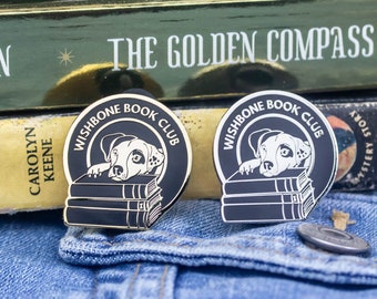 May include: Two gold-colored enamel pins with black and white illustrations of a dog reading a stack of books. The pins feature the text "Wishbone Book Club".