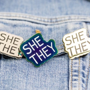May include: Three enamel pins with the text "She They" in white lettering on a blue, gold, and silver background. The pins are attached to a denim jacket.