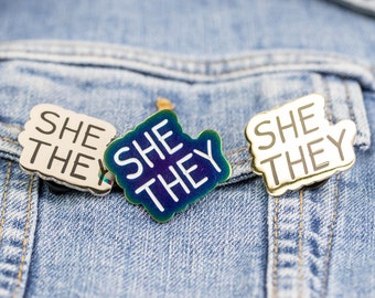 May include: Three enamel pins with the text "She They" in white lettering on a blue, gold, and silver background. The pins are attached to a denim jacket.