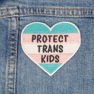 Könnte beinhalten: Ein herzförmiger Aufnäher, der auf eine Jeansjacke genäht ist. Der Aufnäher ist in den Farben der Transgender-Pride-Flagge und trägt den Text "PROTECT TRANS KIDS" in Schwarz.