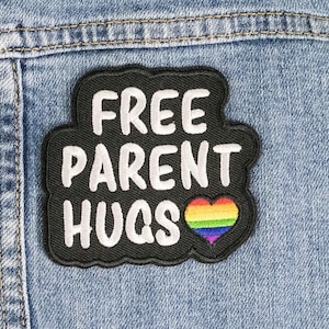 Puede incluir: Un parche negro con texto blanco que dice "FREE PARENT HUGS" y un corazón arcoíris.