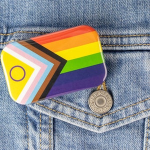 May include: A rainbow pride flag pin with a yellow circle containing a purple ring in the top left corner. The pin is attached to a blue denim jacket.