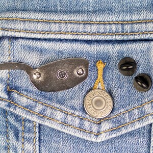 Puede incluir: Un pin de color plateado con forma de gafas de sol, un bot&oacute;n plateado con el texto "Levi Strauss & Co." y dos imperdibles negros en una chaqueta vaquera azul.