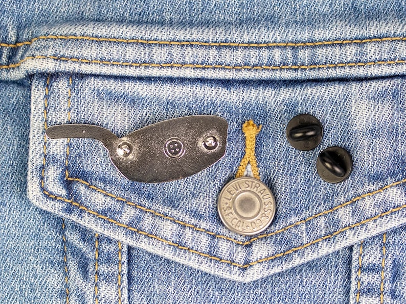 Puede incluir: Un pin de color plateado con forma de gafas de sol, un bot&oacute;n plateado con el texto "Levi Strauss & Co." y dos imperdibles negros en una chaqueta vaquera azul.