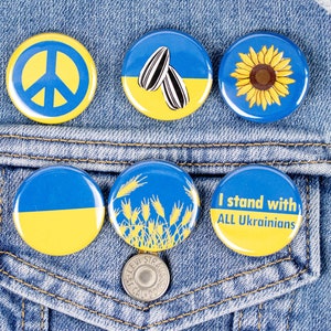 May include: A collection of round pinback buttons with blue and yellow designs, including a peace sign, sunflower, and wheat. One button reads "I stand with ALL Ukrainians." The buttons are displayed on a denim jacket.
