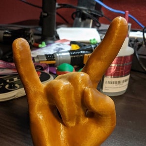Może przedstawiać: Trójwymiarowy model dłoni wydrukowany w kolorze złotym, wykonujący gest rock and roll.