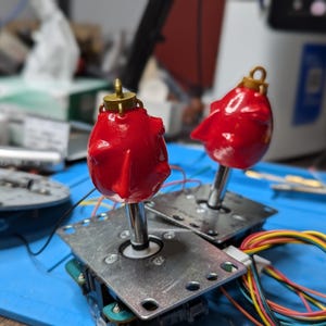 Puede incluir: Dos tapas de joystick de arcade rojas con detalles dorados. Las tapas están montadas en bases plateadas con cables conectados.