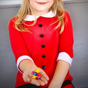 May include: A young girl wearing a red dress with a white collar and black buttons is holding a colorful toy in her hand. The toy is a star-shaped object with multiple colors, including red, yellow, blue, and green.