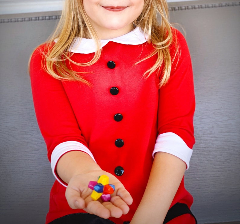May include: A young girl wearing a red dress with a white collar and black buttons is holding a colorful toy in her hand. The toy is a star-shaped object with multiple colors, including red, yellow, blue, and green.