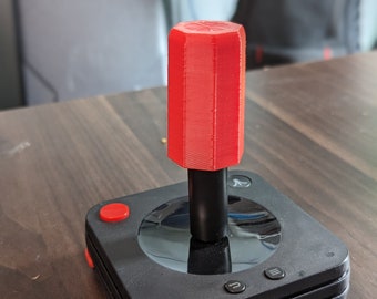 May include: A black Atari joystick with a red top and buttons. The joystick has a black base with a red top and buttons. The joystick is on a dark wood surface.