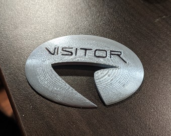 Könnte beinhalten: Ein silberfarbenes, 3D-gedrucktes Abzeichen mit der Aufschrift "VISITOR" in schwarzer Schrift.