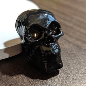 Könnte beinhalten: Eine glänzend schwarze Schädel-Figur mit detaillierten Gesichtszügen. Der Schädel befindet sich auf einer weißen und braunen Oberfläche, was einen auffälligen Kontrast erzeugt. Geeignet für Halloween- oder Gothic-Dekorationen.