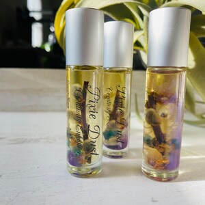 Puede incluir: Tres frascos de vidrio con rodillo y tapas plateadas con etiquetas que dicen "Pixie Dust Creativity Enhancer" y "Pixie Dust Creativity Enhancer". Los frascos están llenos de un líquido transparente y contienen pequeños cristales y flores secas.