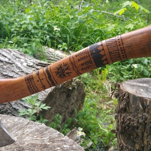 May include: A black-handled axe with a wooden handle that has a decorative burned design. The axe is resting on a tree stump.