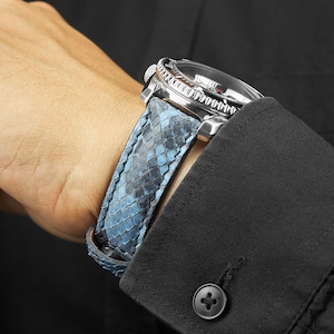 May include: A wristwatch with a blue snakeskin-effect leather strap. The watch has a silver case and a black face. The watch is worn on a wrist, with a black shirt sleeve in the background. The strap is patterned.