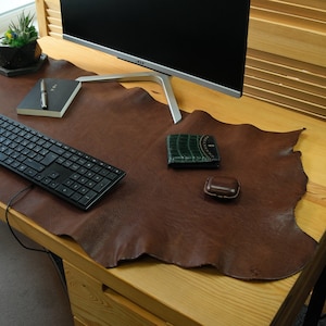 Puede incluir: Una alfombrilla de escritorio de cuero marrón con un teclado negro, una cartera verde y una funda de cuero marrón sobre un escritorio de madera con un monitor de ordenador negro.
