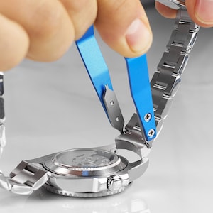 May include: A silver-toned metal watch with a link bracelet being adjusted by a bright blue spring bar tool. The tool features two metal prongs that are inserted into the watch lugs to detach the bracelet from the watch case.