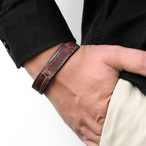 Puede incluir: Un brazalete de cuero marrón con un patrón de cocodrilo.