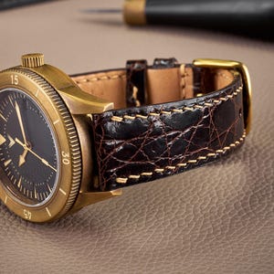 Könnte beinhalten: Eine Armbanduhr mit einem runden, bronzefarbenen Gehäuse, einem schwarzen Zifferblatt und einem dunkelbraunen, strukturierten Lederarmband mit hellen Nähten. Die Uhr verfügt über eine drehbare Lünette mit eingravierten Zahlen und eine bronzefarbene Schließe.