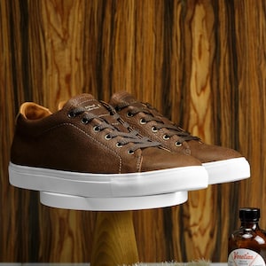 Könnte beinhalten: Braune Lederschuhe mit weißen Sohlen, eine Holzschuhbürste, eine Flasche neutrale Schuhcreme und ein Glas schwarzer Schuhpolitur. Die Produkte sind auf einer weißen Oberfläche mit einem Holzhintergrund angeordnet.