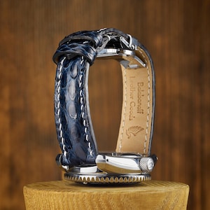 May include: A wristwatch featuring a dark blue textured leather strap with white stitching. The interior of the strap is light tan leather with the text Baldanoff Leather Goods and a small logo embossed on the surface. The watch has a silver metal case.