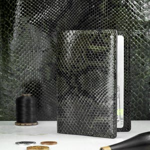 Pode incluir: Uma carteira de couro texturizado verde escuro com um padrão de pele de cobra. A carteira está aberta, revelando um cartão. Uma bobina de linha preta, moedas e uma ferramenta também são visíveis.