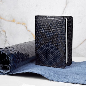 Könnte beinhalten: Ein dunkelblaues, strukturiertes Lederportemonnaie steht aufrecht neben einem gerollten Stück passenden Leders. Das Portemonnaie hat ein reptilienartiges Muster. Der Hintergrund ist eine weiße Marmoroberfläche.