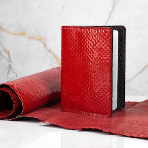 Könnte beinhalten: Eine rote Lederbörse mit Schlangenhautmuster. Die Börse ist geschlossen und hat einen schwarzen Rand. Die Börse liegt auf einer weißen Marmorfläche.